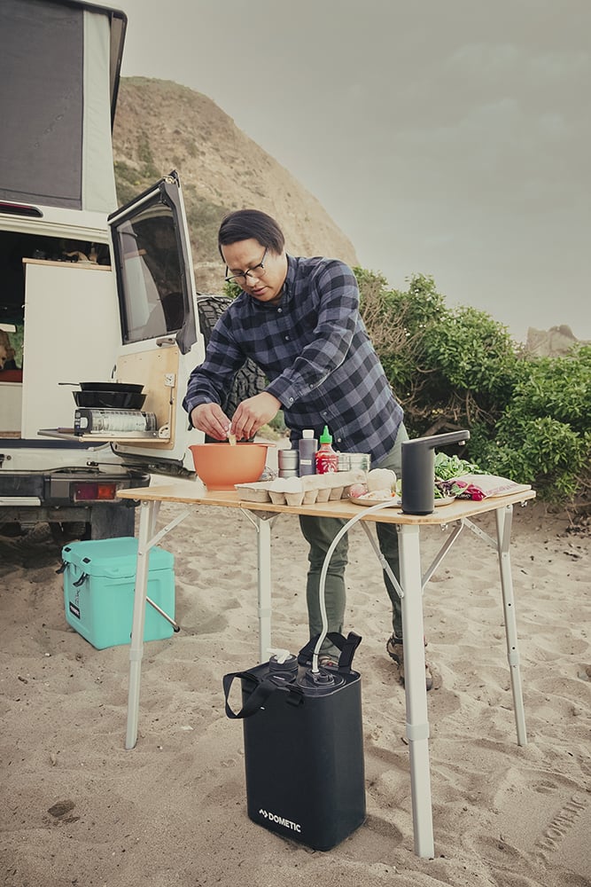 A man preparing food outdoors on a Dometic table with a Dometic water jug and cooler.