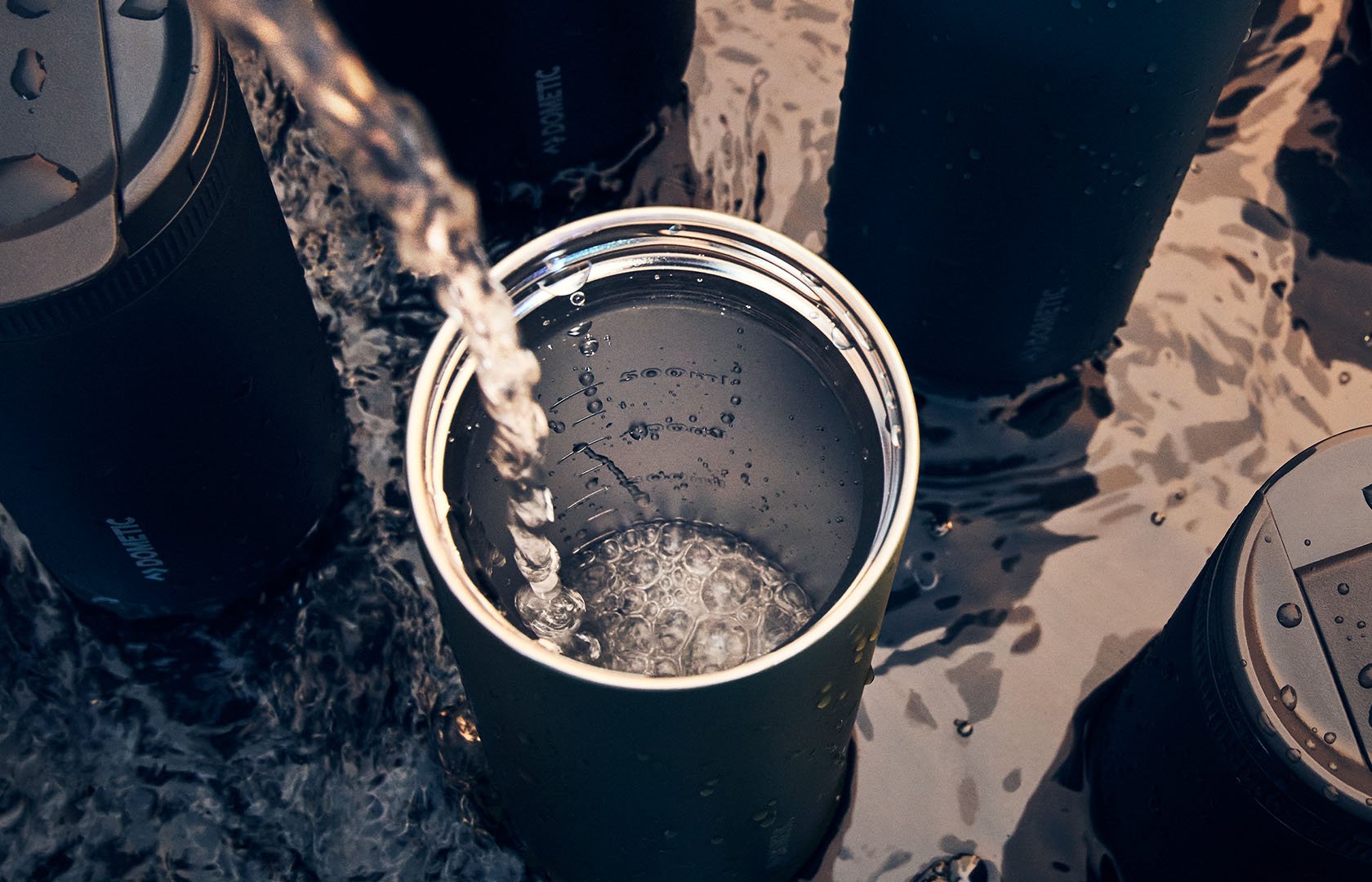 Isolerad metallmugg som fylls med vatten i grunt vatten utomhus, omgiven av andra dryckesbehållare.