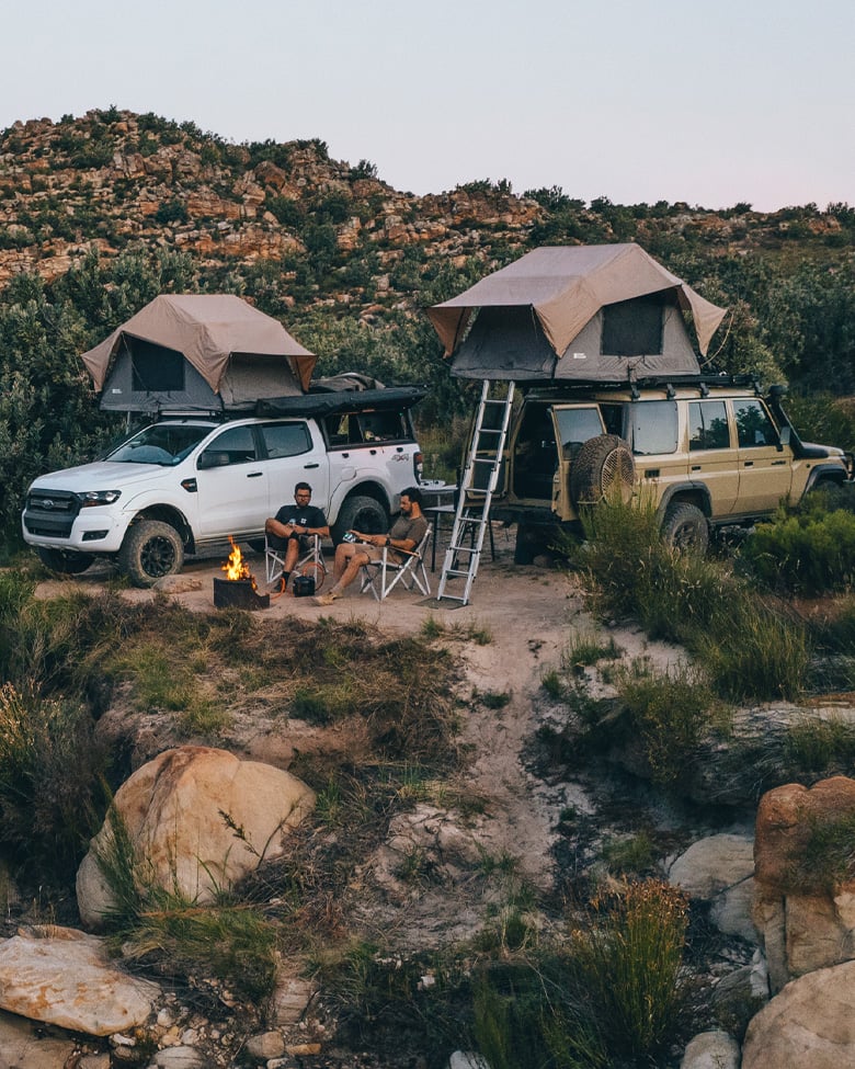 Two off-road vehicles with rooftop tents parked in rocky landscape, camping setup with chairs and campfire in outdoor setting