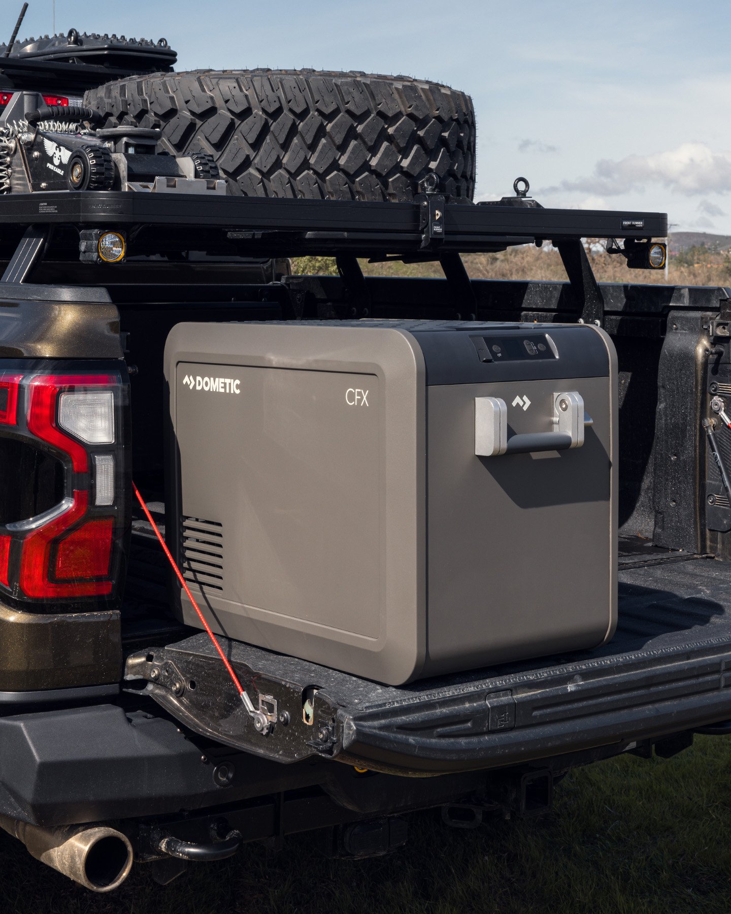 Dometic CFX powered cooler placed on the open tailgate of a pickup truck with an off‑road spare tire mounted on the rack above in an outdoor setting.