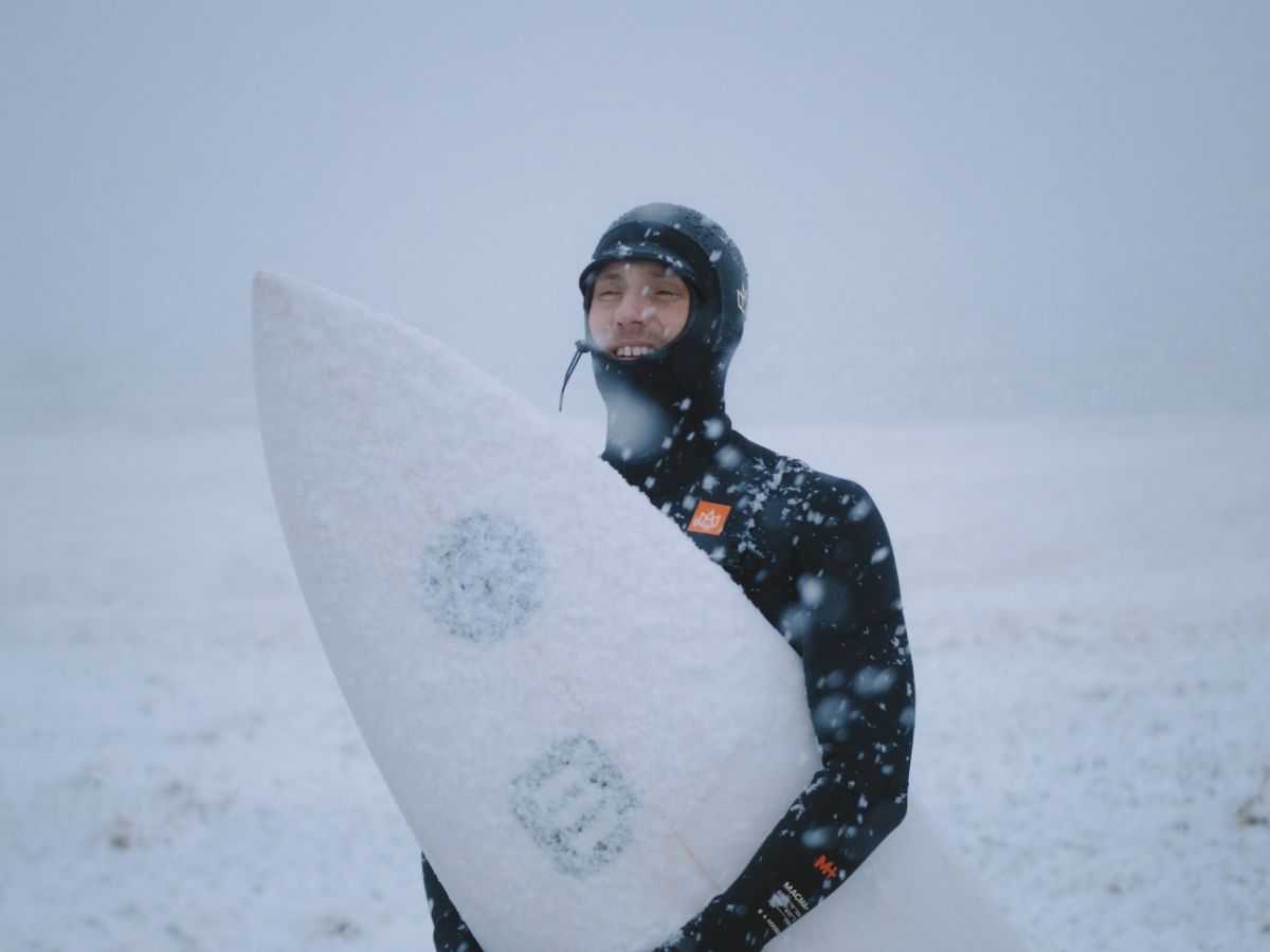 A surfer dressed in a full wetsuit in a snowy landscape, smiling while holding a surfboard and covered in fresh snow.