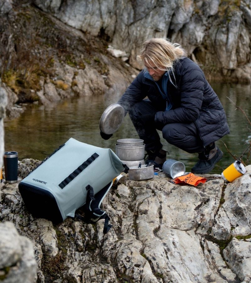 A person crouched on rocks by a river preparing a meal and surrounded by outdoor gear