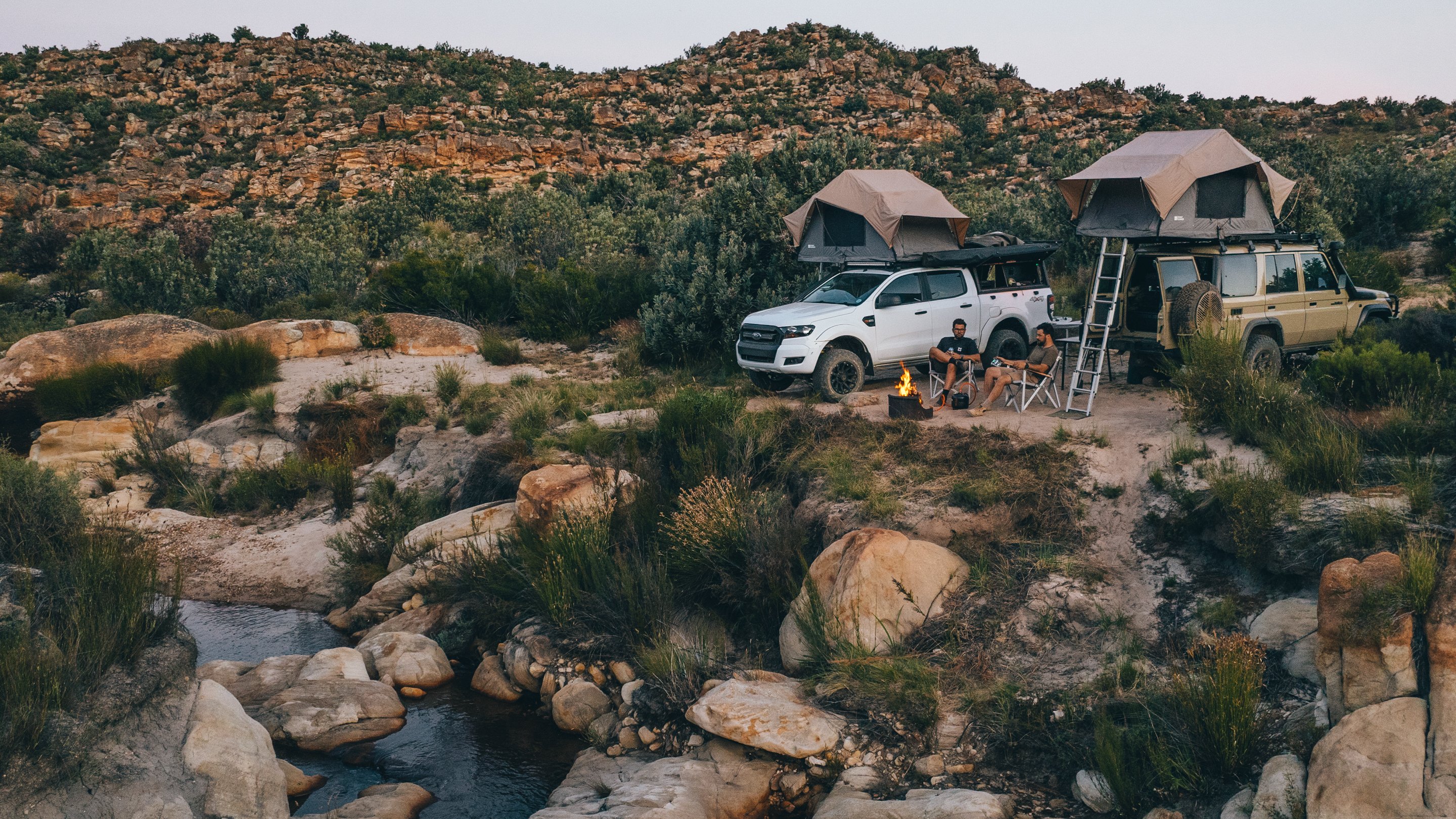 Two off-road vehicles with rooftop tents parked in rocky landscape, camping setup with chairs and campfire in outdoor setting