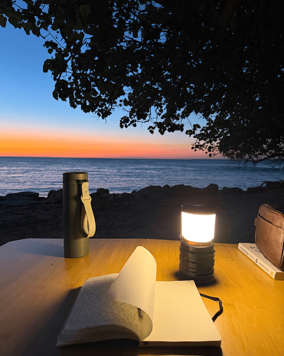 Harry Holland sits at his Dometic bamboo camp table as the sun sets over Stradbroke Island. Lit by a camp light, he journals about an incredible first day of camping and adventure, creating lasting memories while enjoying life in the great outdoors.
