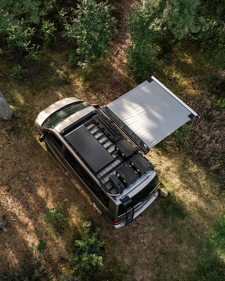 Draufsicht auf einen Campervan mit Dachträgersystem und ausgefahrener Seitenmarkise in einer Waldlichtung.