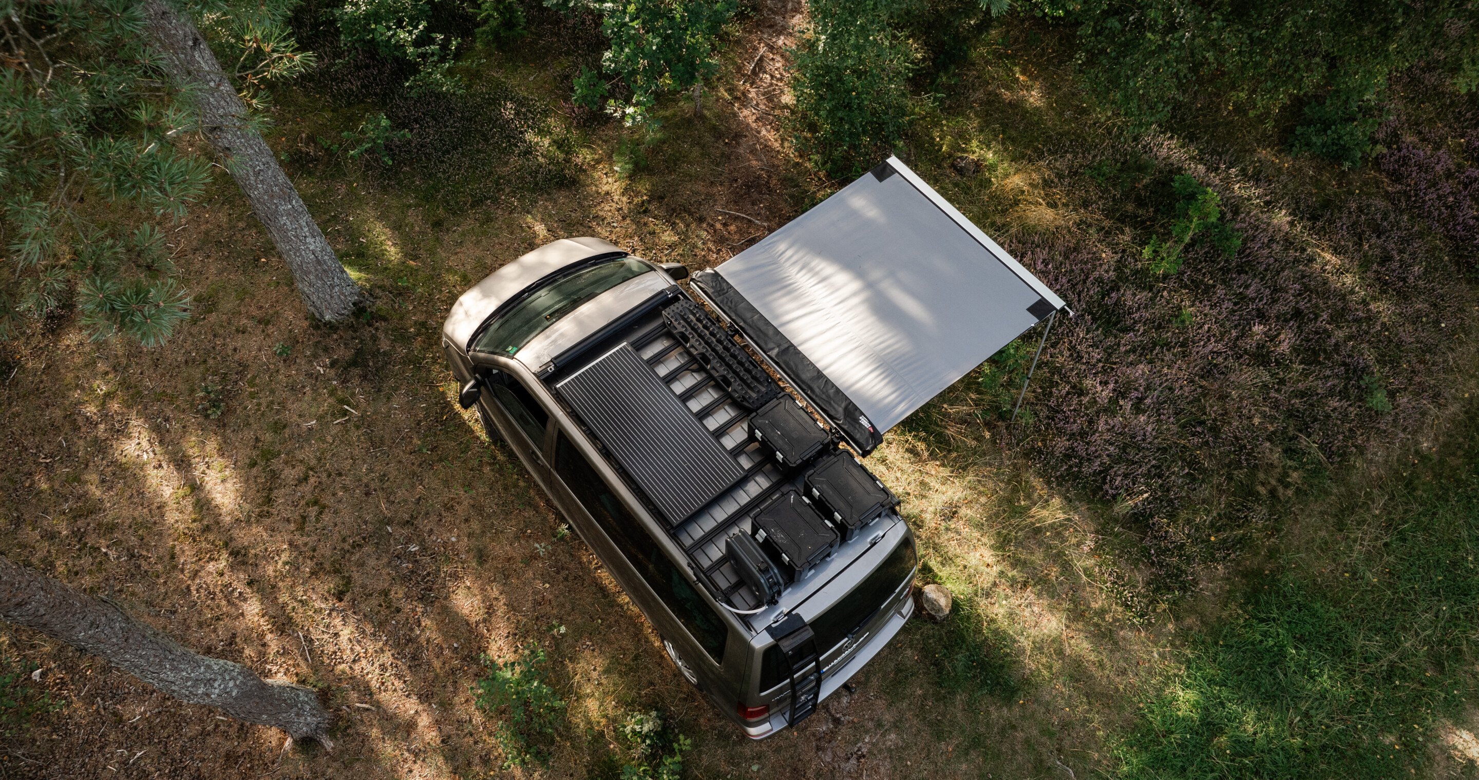 Vue aérienne d’un fourgon équipé d’une galerie de toit et d’un auvent latéral déployé dans une clairière forestière.