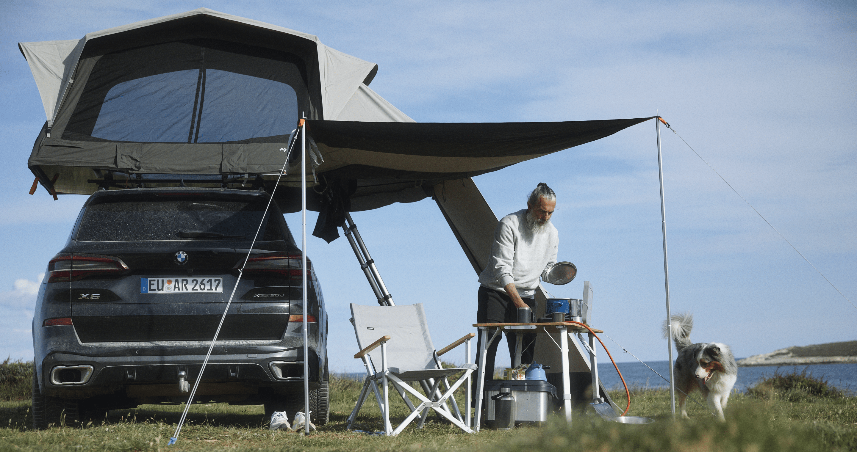 Man and dog in a rooftop tent and awning setup with camping furniture in lush, summery surroundings