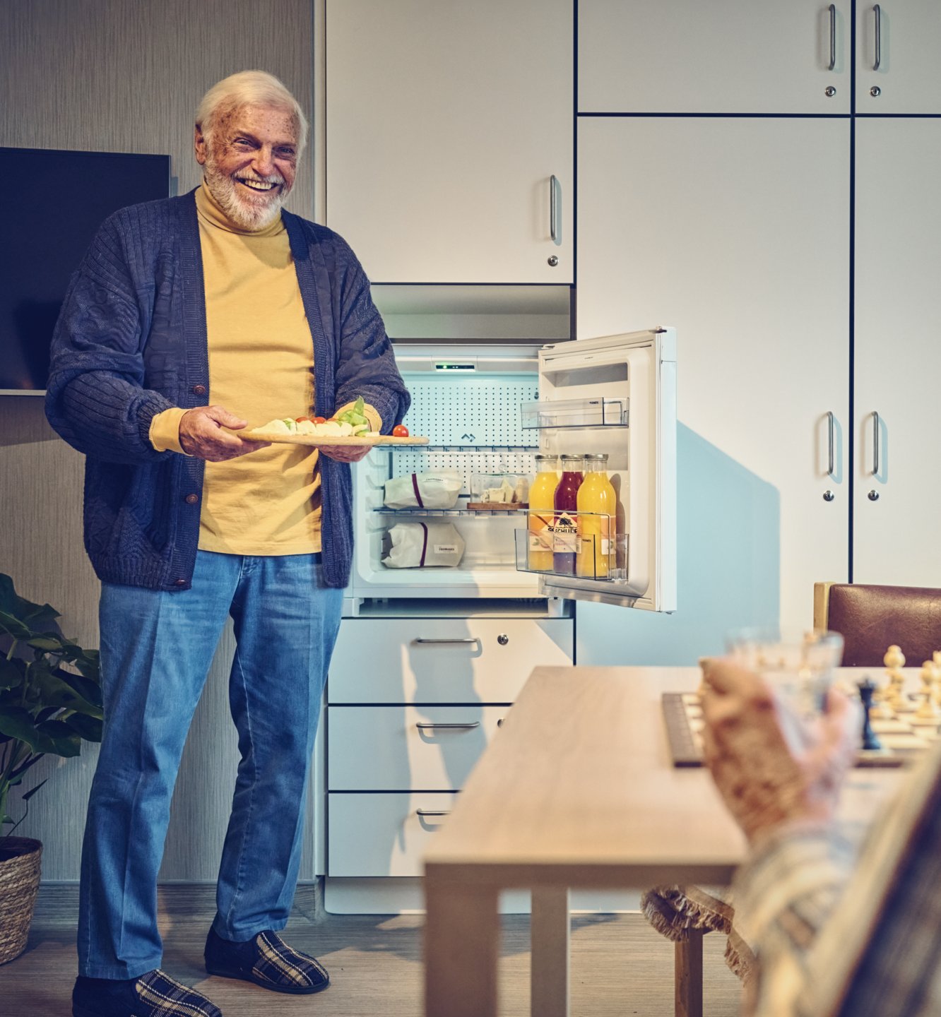 Elderly man offering a plate of food from a minifridge in a care facility room.