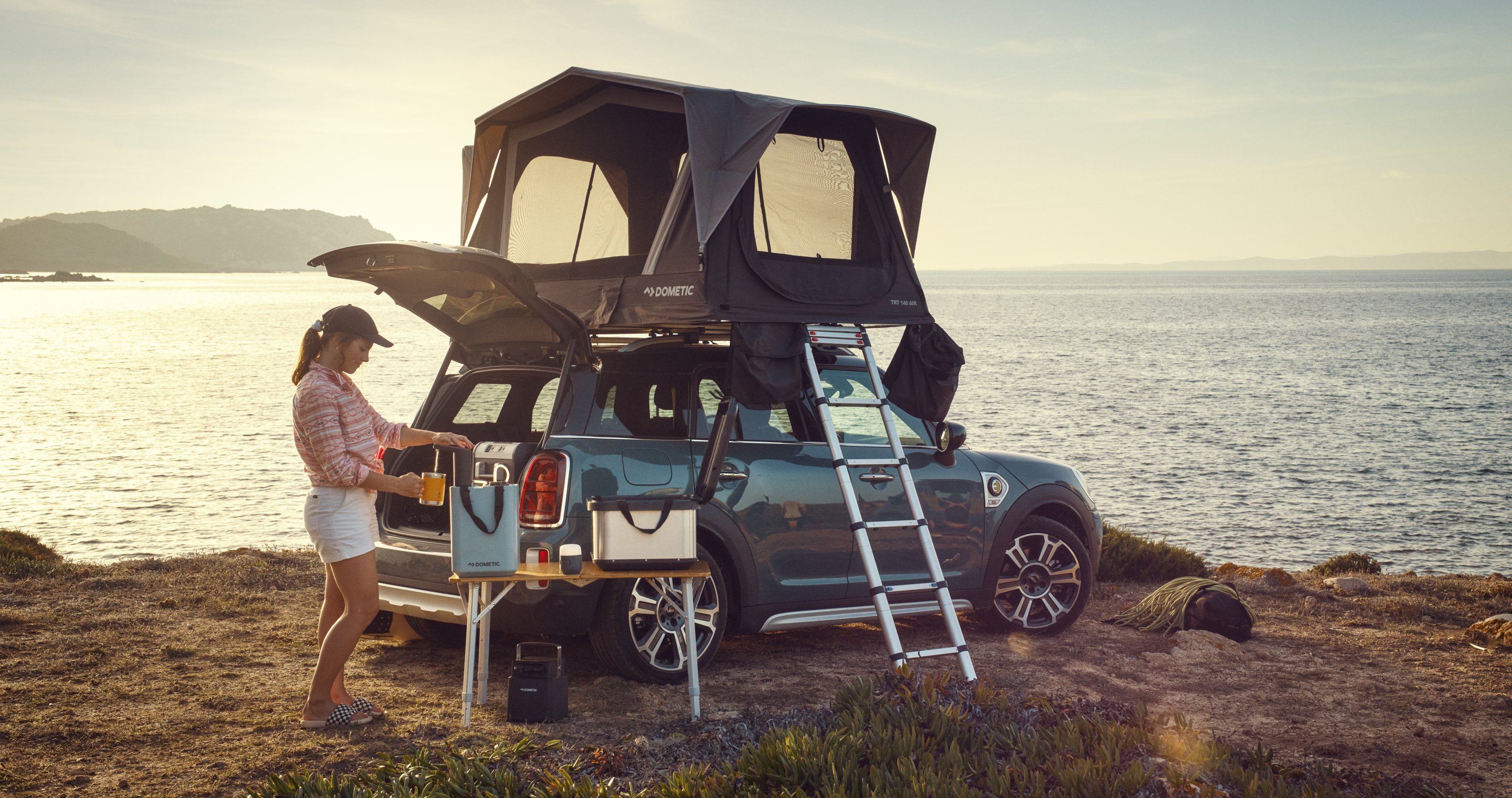 Woman setting up camp with a Dometic rooftop tent mounted on a compact SUV, parked by the ocean at sunset — ideal for rooftop tent travel guides and outdoor adventure content