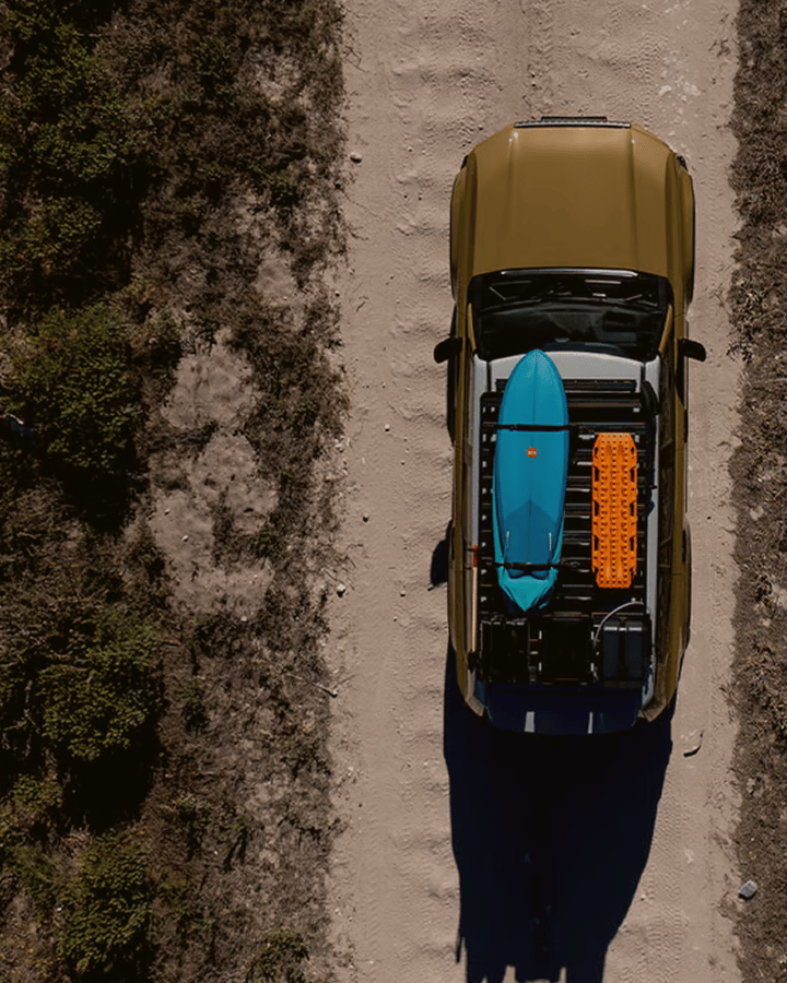 Vue aérienne verticale d’un véhicule sur une piste sablonneuse avec une galerie de toit transportant une planche de surf et une planche de traction.