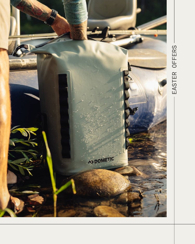 Dometic waterdichte rugzak die in ondiep water naast stenen en planten wordt neergezet, met een kajak op de achtergrond in een buitensituatie aan het water.