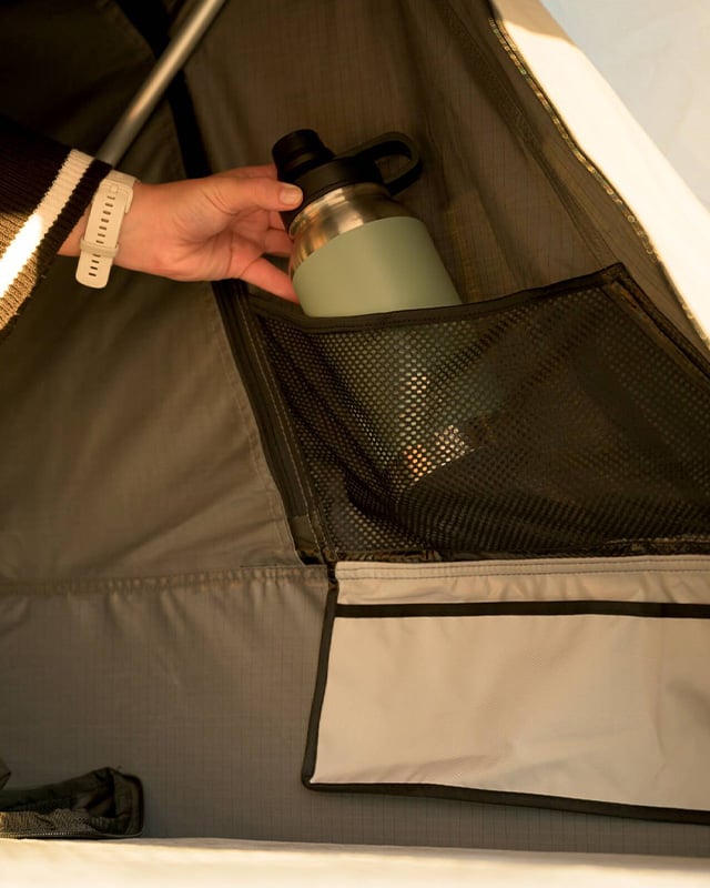 Mano che ripone una borraccia isolata verde nella tasca in rete all’interno della tenda da tetto Front Runner Dometic Feather‑Lite II.
