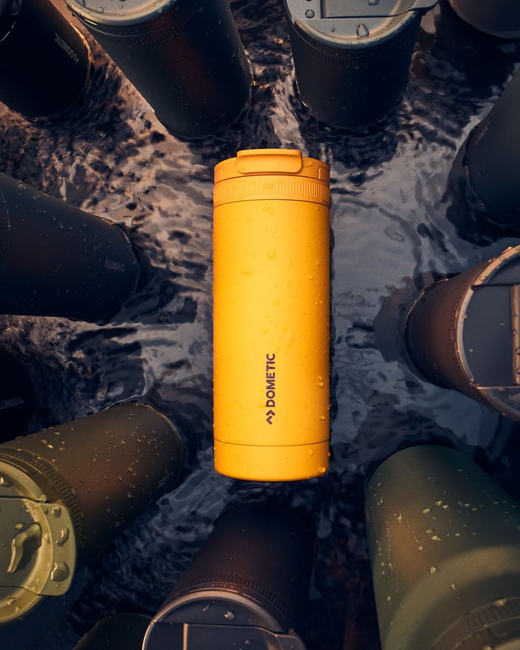 Gul isolerad termomugg med vattendroppar omgiven av utomhusmuggar i grunt vatten.