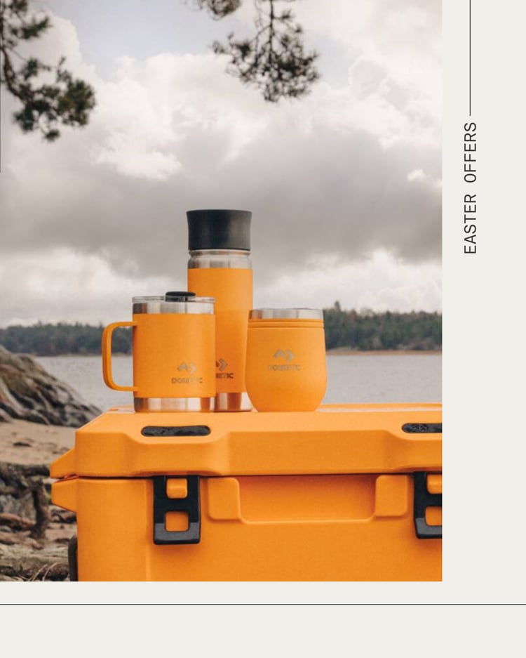 Orange Dometic-kylbox med matchande isolerad mugg, tumbler och kopp, placerad utomhus vid en sjö under molnig himmel.